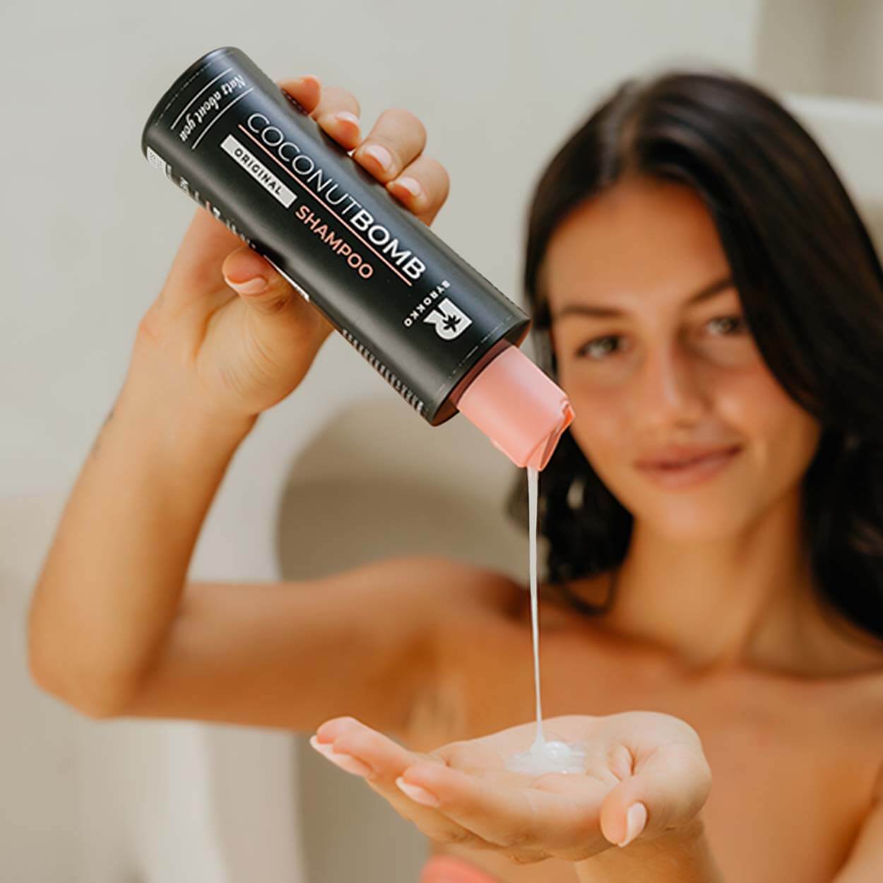 A brunette girl is pouring Byrokko Coconut Bomb shampoo on her hand.